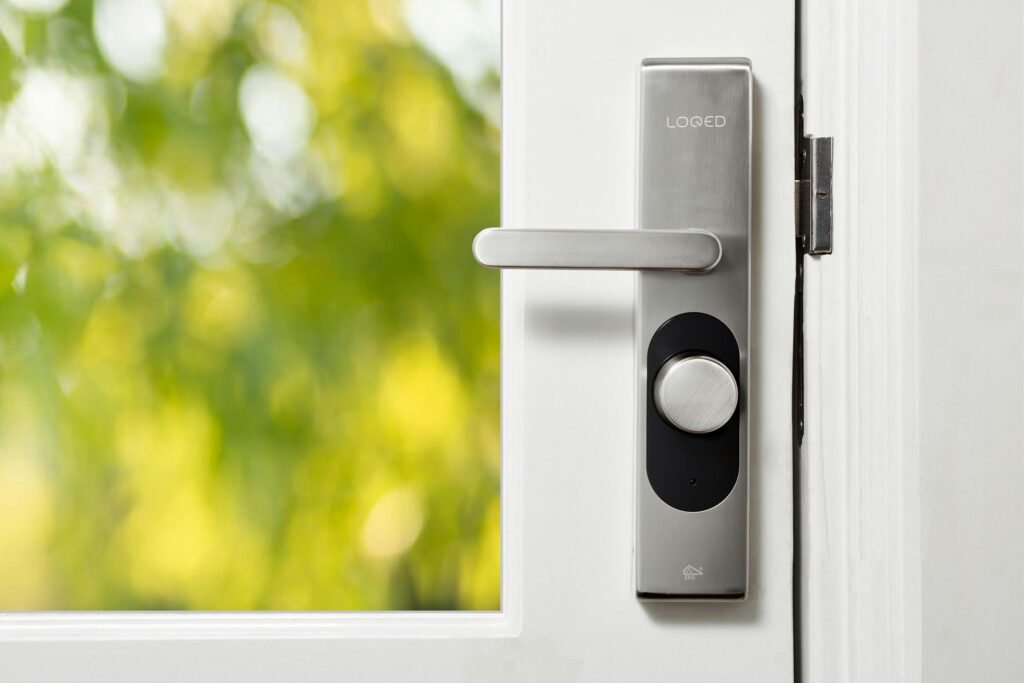pexels photo 6689289 6689289 Sleek smart door lock on a white door with blurred green foliage in the background.